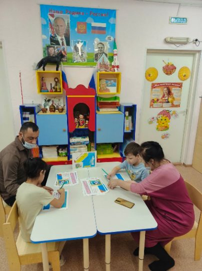 Фото 3. Участники викторины «Что мы знаем о своей стране и выборах», проведенной в рамках Дня молодого избирателя в  детском саду  № 61 «Лель» отвечают на вопросы викторины. Дети отвечают на вопросы вместе с родителями. Сидят за столами и отмечают ответы  на вопросы викторины на распечатанных бумажных листах. 