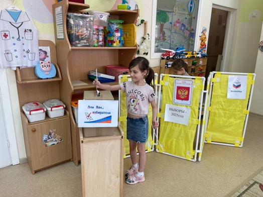 Фото 2. Участники Дня молодого избирателя в детском саду №18 «Мишутка» принимают участие в сюжетно-ролевой игре: Выборы «Президента России». Избиратель опускает свою бюллетень в ящик для голосования. 
